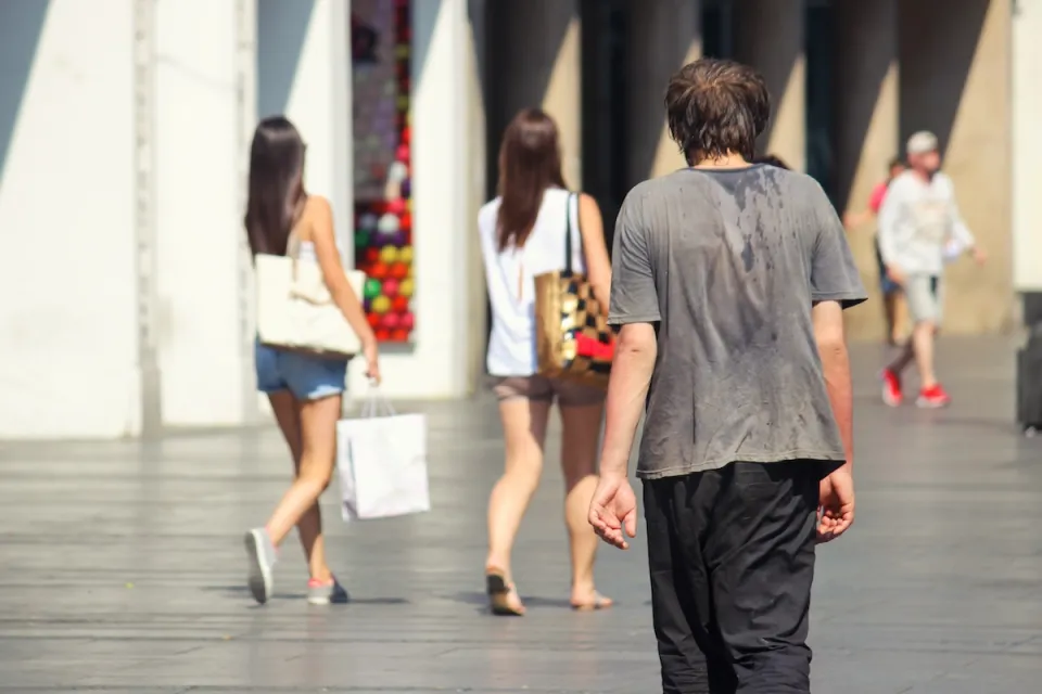 Man walking down city sidewalk with sweat on back of his t-shirt on hot day.